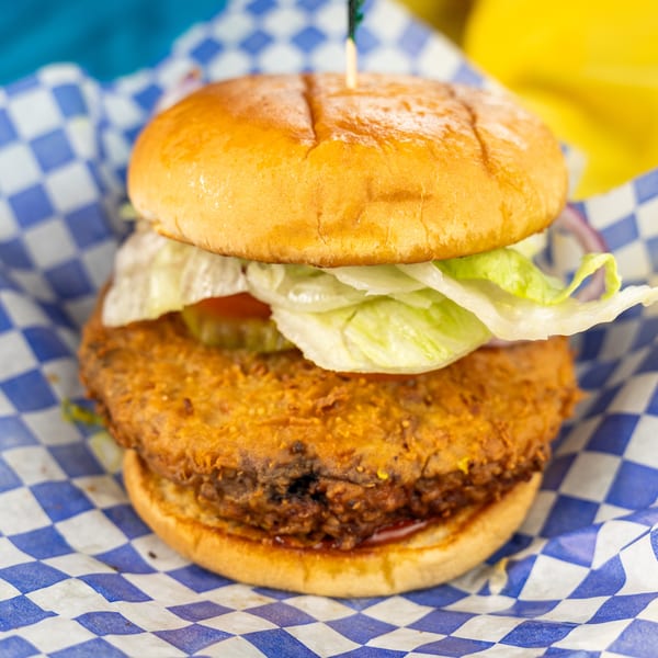 Crispy fried chicken sandwich with lettuce, tomato, and pickles on a bun in checkered paper.