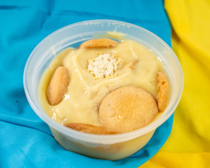 Banana pudding in a clear plastic container with vanilla wafers and crumbled topping.