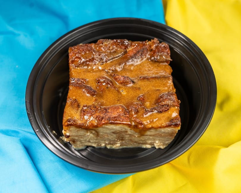 A square of bread pudding with a glossy caramel topping in a black bowl on a blue and yellow cloth.