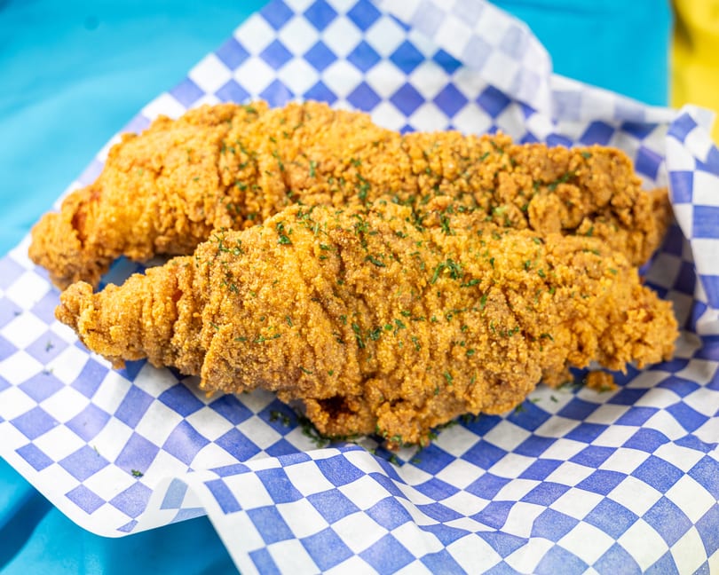 Two golden-brown fried fish fillets, sprinkled with parsley, resting in a blue and white checkered paper basket.