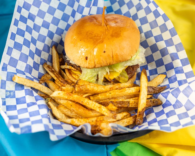 Cheeseburger with lettuce and pickles, served with seasoned fries in a blue and white checkered basket.