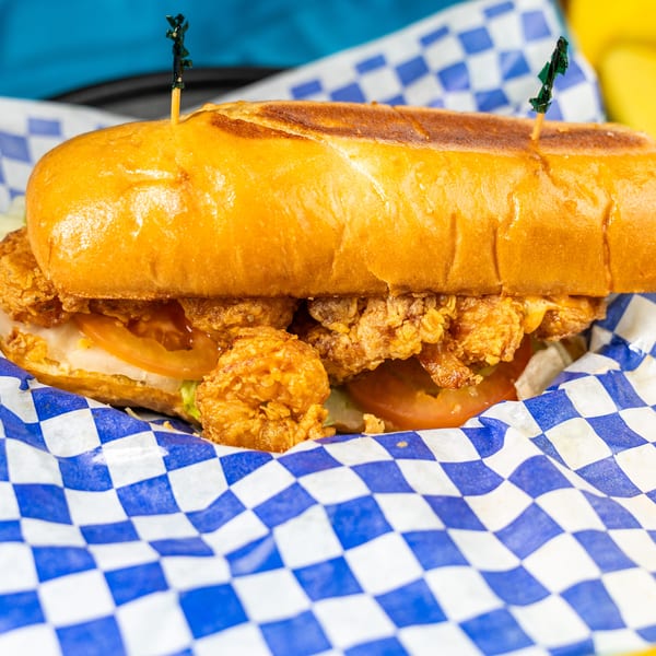 Close-up of a fried shrimp po'boy sandwich with lettuce and tomato in blue and white checkered paper.