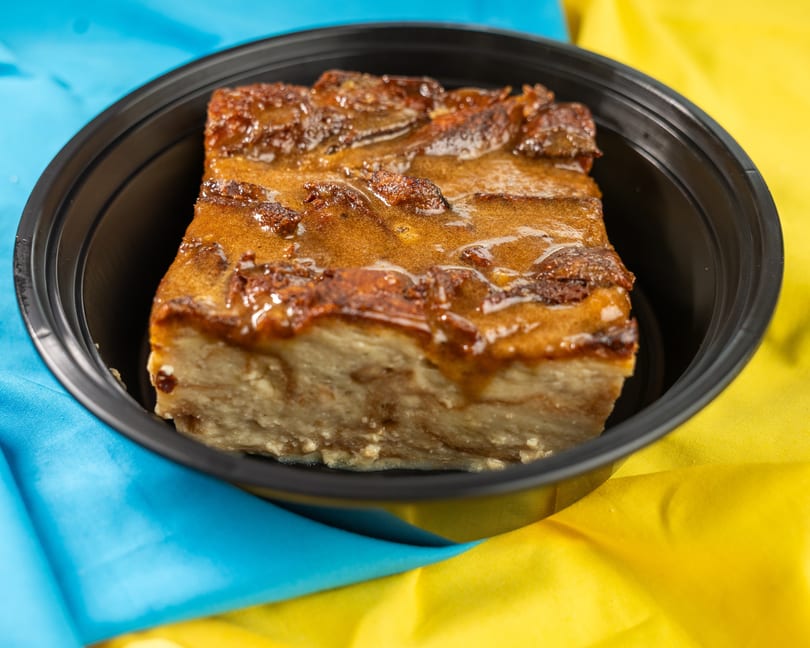 Bread pudding with caramel sauce in a black container, on a blue and yellow tablecloth.